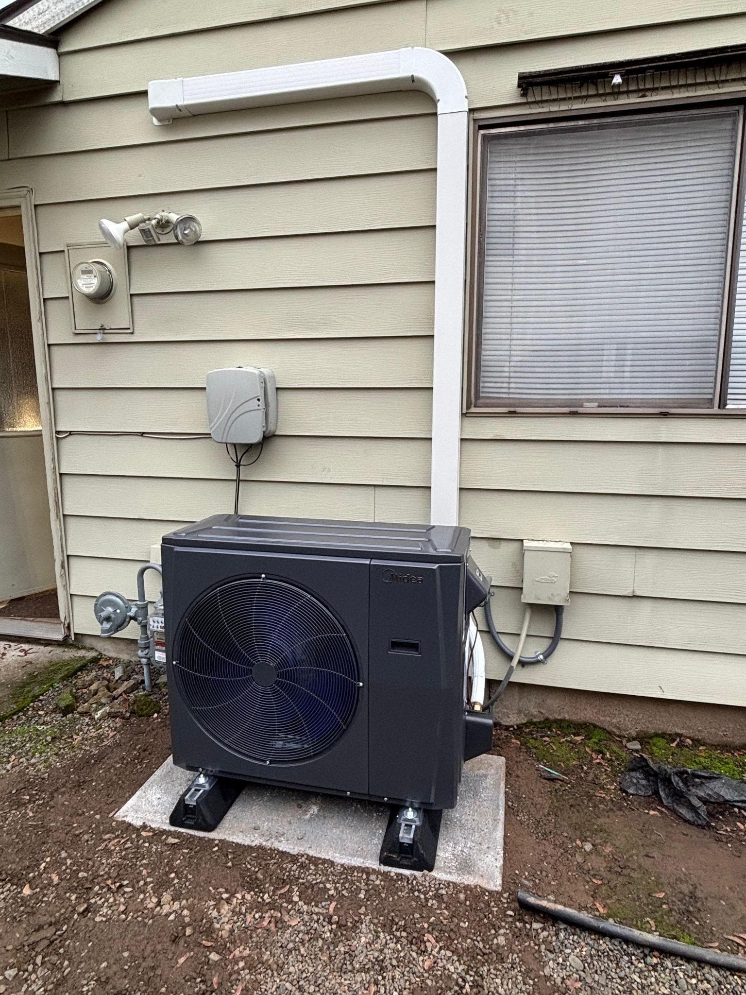 Midea cold-climate heat pump installed on raised supports beside a Tigard home, with a covered line set running along the siding.